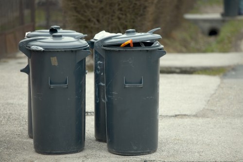 Two large, dark grey plastic wheelie bins with lid handles are positioned side by side on a paved driveway. The bin on the left has its lid closed, showing a smooth, slightly textured surface with a small rectangular label area, while the bin on the right has its lid partly open, revealing an orange-handled tool or object resting inside. The bins are situated outdoors, with a background of blurred trees and a concrete bridge or structure, indicating a residential or suburban setting. The environment appears to be well-lit, with natural daylight illuminating the scene. Flat Clearance Twickenham offers rubbish removal services, and these bins are typical of waste collection practices, often used for general household waste. The setting suggests regular waste disposal activities, with the bins ready for collection within a property in Twickenham, part of the London borough. This visual context aligns with promoting effective waste management and recycling efforts in the local area.
