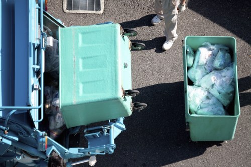 An overhead view of a light green, large plastic rubbish bin positioned on a dark asphalt surface in an outdoor setting, partially open with a collection of transparent plastic bags filled with mixed waste visible inside. Adjacent to the bin is a blue bin loading vehicle, which is partially visible on the left side of the image and appears to be used for rubbish collection. A person dressed in light-colored clothing and white footwear is standing nearby, with only their legs and feet visible in the image. The background shows a section of the pavement and a small outdoor air conditioning unit mounted on a nearby wall, suggesting a residential or small commercial area. This scene illustrates typical rubbish collection activity that a company like Flat Clearance Twickenham might facilitate in the local area for efficient waste removal, including recyclable and general waste disposal while maintaining a tidy environment in Twickenham or surrounding boroughs.