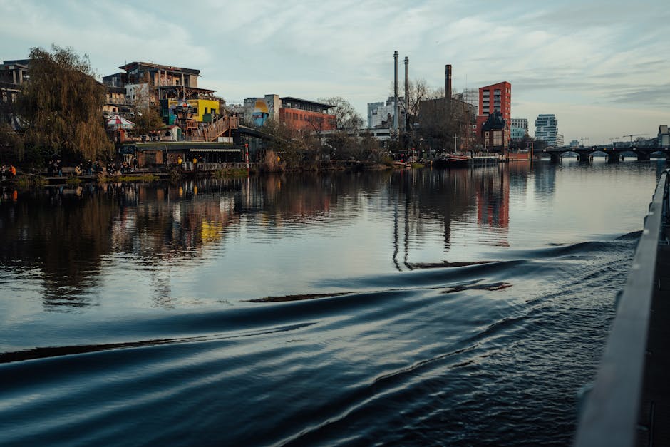 The image depicts a calm river meandering through an urban landscape, with buildings and industrial structures along the riverbank. On the left side, there are multi-story buildings, including a yello