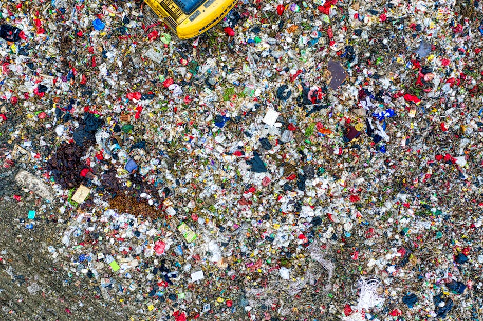 An extensive aerial view of a large landfill site filled with a dense layer of mixed waste materials, including plastic wrappers, cardboard pieces, broken textiles, and other household waste. The surf